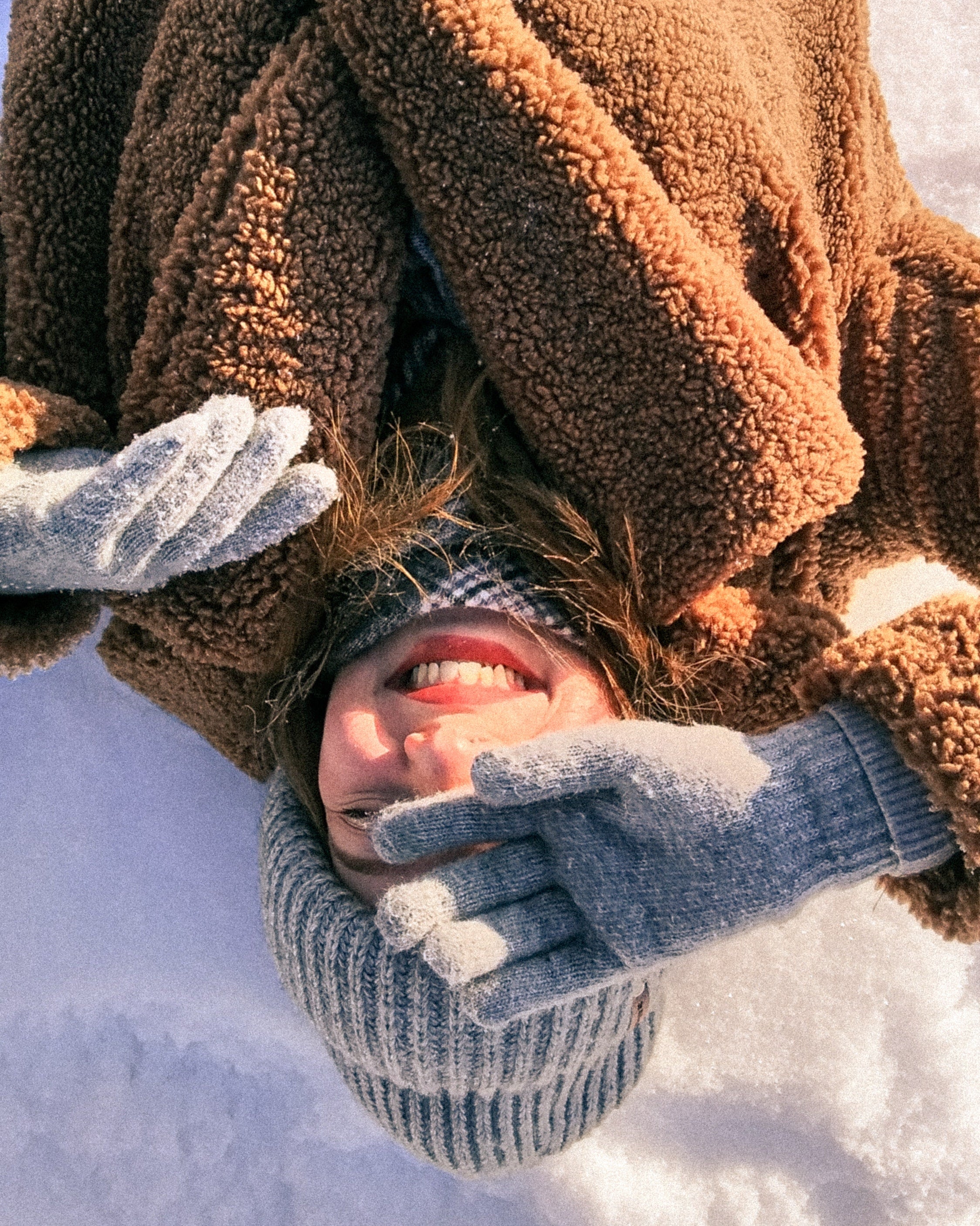 A woman in a beanie and gloves laying in the snow under the sun